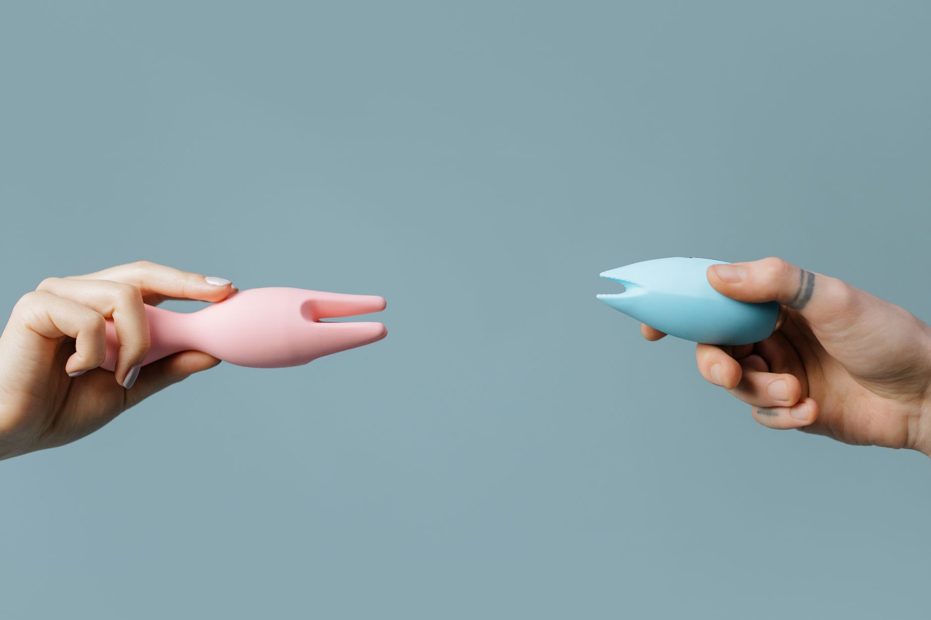 Two hands holding clitoral vibrators against a pastel background, showcasing external pleasure tools